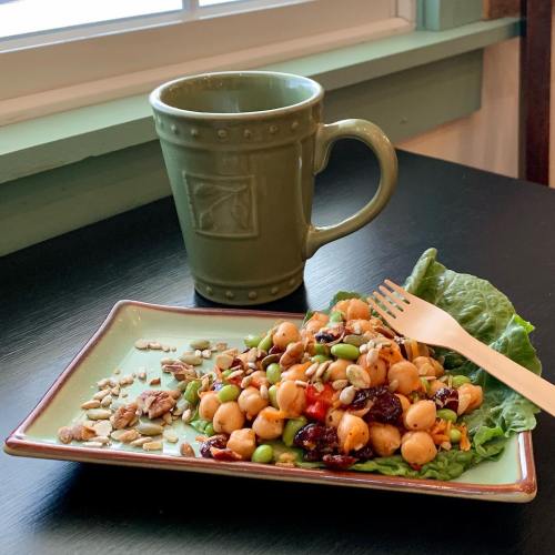 A colorful salad with chickpeas, edamame, and seeds on a plate, next to a green mug on a black table.
