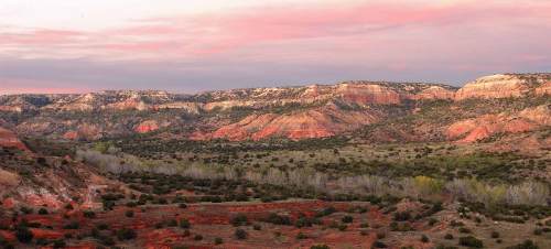 Vast landscape of colorful rock formations and rolling hills under a pink and purple sky at sunset.