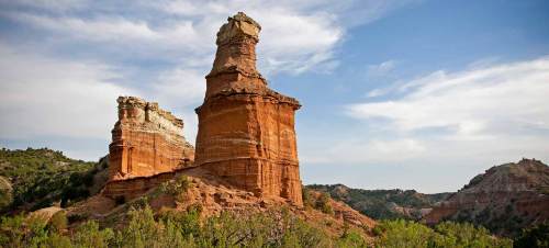 A towering rock formation with layered red and white cliffs under a blue sky, surrounded by green vegetation.