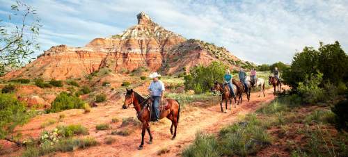 A group of riders on horseback traverses a scenic trail with colorful rock formations in the background.