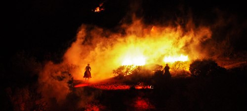 A silhouette of a person on horseback rides through a fiery landscape, surrounded by smoke and flames at night.