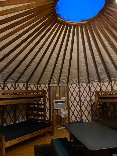 Interior of a yurt with wooden beams, bunk beds, a table, and a circular skylight revealing a blue sky.