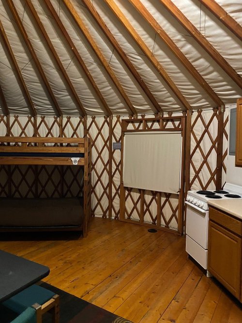 Interior of a yurt featuring wooden beams, a bunk bed, a kitchenette, and a wooden floor.