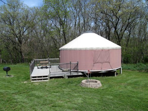 A pink yurt on a wooden deck surrounded by trees, with a grill and fire pit in a grassy area.