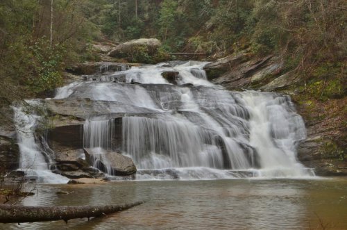 A serene waterfall cascading over rocky ledges into a calm pool, surrounded by lush greenery and trees.