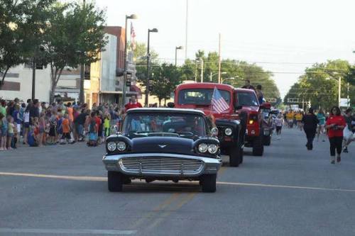 A classic black car with an American flag leads a parade, surrounded by red trucks and a crowd of spectators.