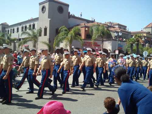 A military parade featuring uniformed personnel marching down a street, with spectators and palm trees in the background.