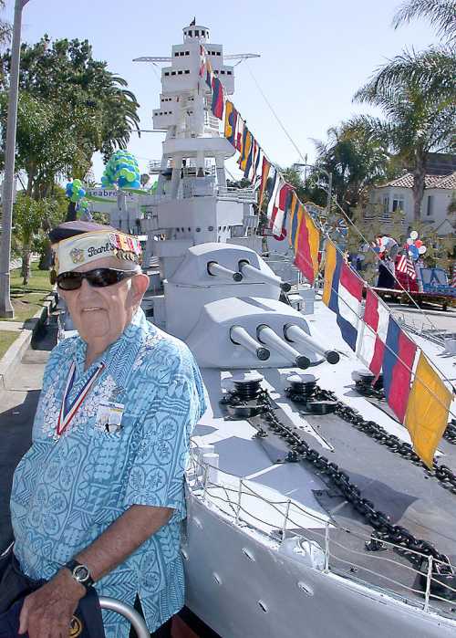 An elderly man in a blue shirt stands beside a large battleship adorned with colorful flags at a festive event.