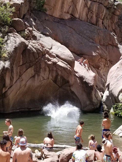 A group of people by a rocky riverbank, with one person jumping into the water, creating a splash.