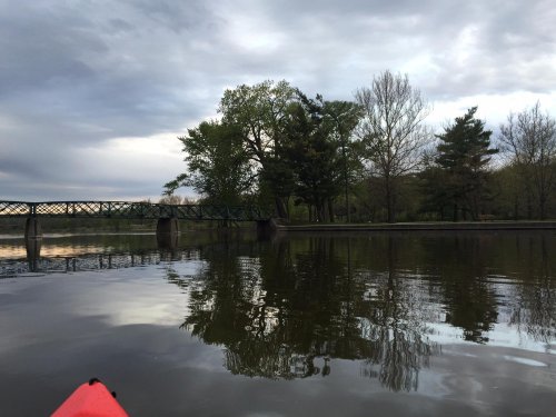 A kayak's red bow in the foreground, reflecting trees and a bridge on a calm, cloudy lake.