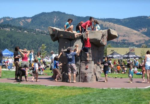 Children and adults climbing a rock wall in a park, with a crowd and mountains in the background on a sunny day.