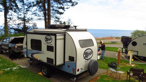 A small camper trailer parked near the ocean, with people setting up a campsite and trees in the background.