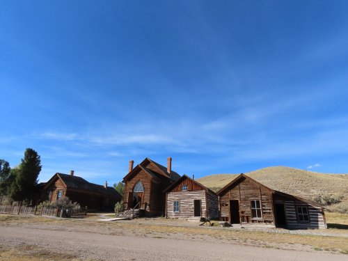 Abandoned wooden buildings under a clear blue sky, set against a backdrop of rolling hills.