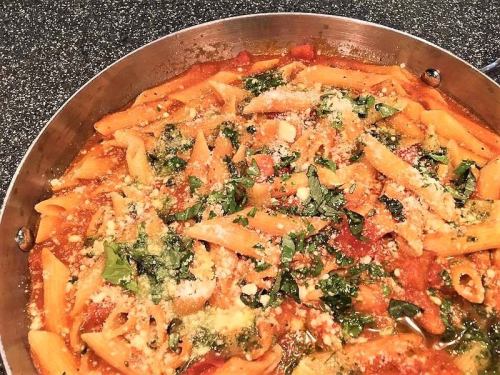 A close-up of penne pasta in a sauce with tomatoes, spinach, and cheese, served in a stainless steel pan.