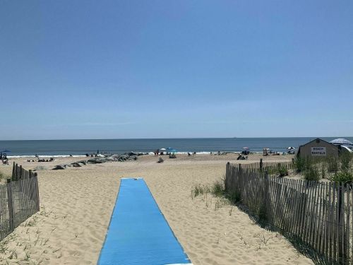 A sandy beach with a blue walkway leading to the ocean, surrounded by umbrellas and beachgoers under a clear sky.