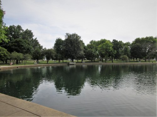 A serene park scene featuring a calm pond surrounded by lush green trees and benches along the water's edge.