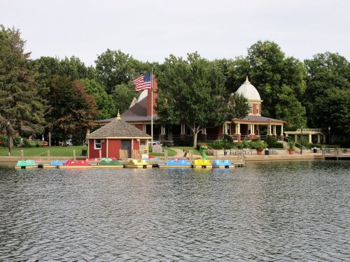 A serene lake scene featuring colorful paddle boats, a red boathouse, and a historic building with an American flag.