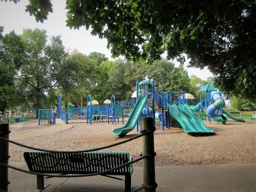 A playground with blue slides and climbing structures, surrounded by trees and a bench in the foreground.