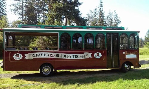 A red and green trolley bus with large windows, labeled "Friday Harbor Jolly Trolley," parked in a grassy area.