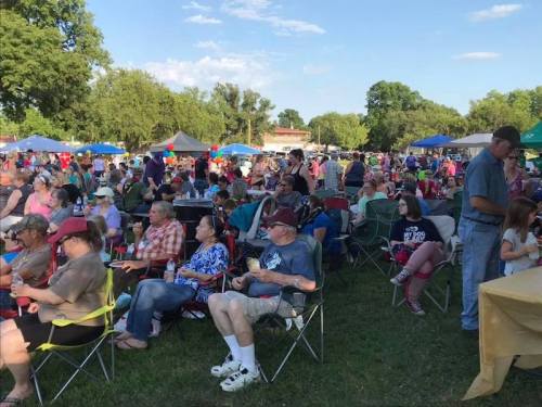 A large crowd enjoys an outdoor event, sitting in chairs under blue skies with tents and trees in the background.