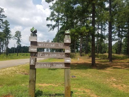 Wooden sign welcoming visitors to Paul M. Grist State Park, surrounded by tall pine trees and a clear blue sky.