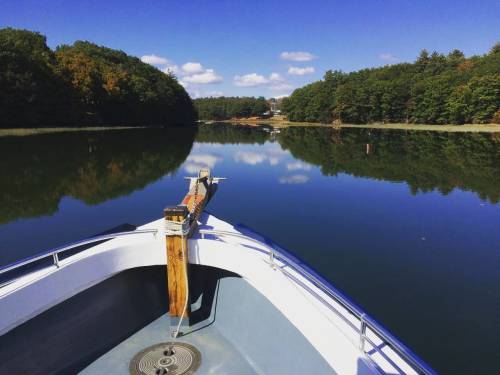 A boat's bow on calm water, surrounded by trees and a clear blue sky, reflecting the serene landscape.