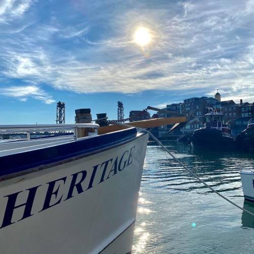 A boat named "Heritage" docked in a harbor under a bright sun and blue sky, with buildings in the background.