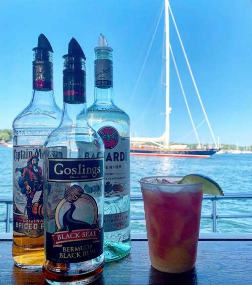 Three bottles of rum and a cocktail with a lime slice, set against a sunny waterfront with a sailboat in the background.