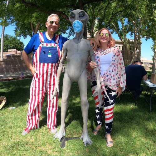 A man in striped overalls and a woman in patriotic attire pose with a gray alien statue wearing a mask in a park.