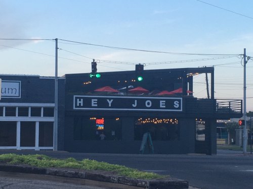 Exterior of a bar named "Hey Joes," featuring green traffic lights and red umbrellas on the patio.