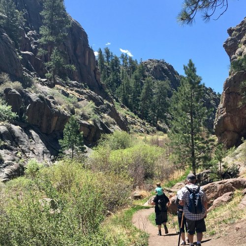 Two hikers on a trail surrounded by rocky cliffs and lush greenery under a clear blue sky.