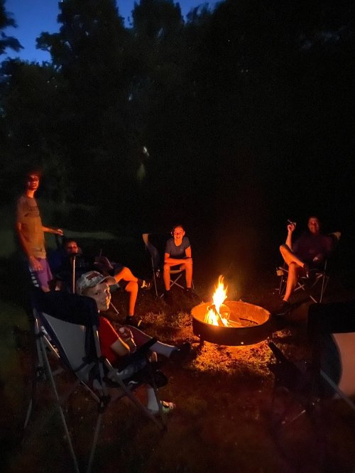 A group of people sitting around a campfire at night, enjoying each other's company in a dark outdoor setting.