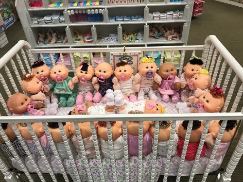 A crib filled with various baby dolls, each dressed differently and some with pacifiers, arranged in a playful display.