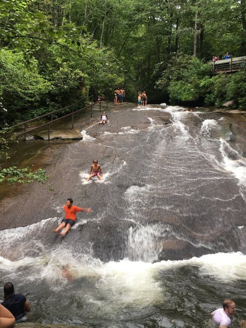 People sliding down a smooth, rocky water slide in a forested area, with others enjoying the water nearby.
