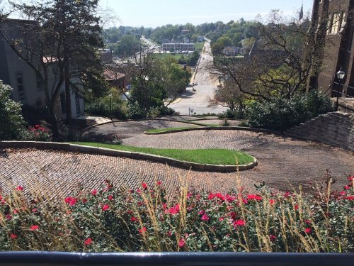 A winding cobblestone path lined with flowers descends towards a street in a scenic, hilly area.