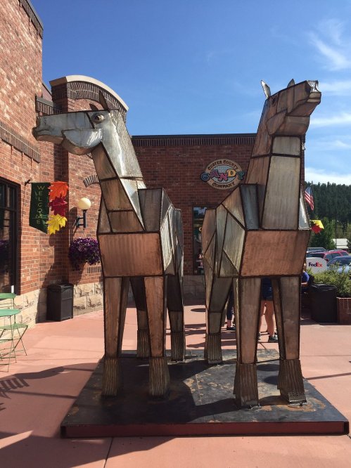 Two large metal horse sculptures stand outside a brick building, surrounded by flowers and flags.