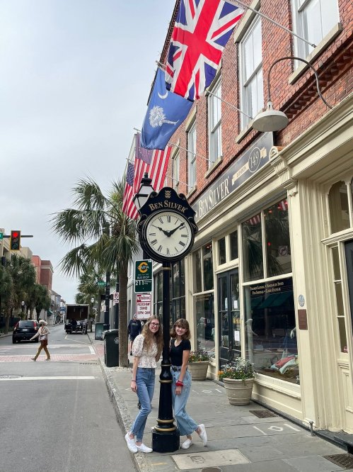 Two young women stand by a vintage clock on a street lined with flags and historic buildings.