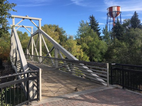 A metal bridge spans a small river, with trees in the background and a water tower visible nearby under a blue sky.