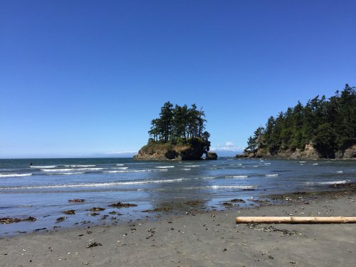 A serene beach scene with gentle waves, a small island with trees, and a clear blue sky in the background.