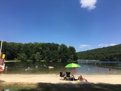 A sandy beach by a calm lake, with people swimming and relaxing under a green umbrella on a sunny day.