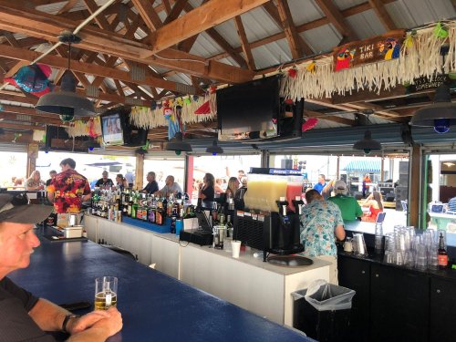 A lively tiki bar scene with patrons enjoying drinks, colorful decor, and a bustling atmosphere under a wooden roof.