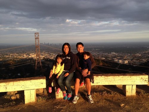 A family of four sits on a bench overlooking a city skyline at sunset, with mountains in the background.