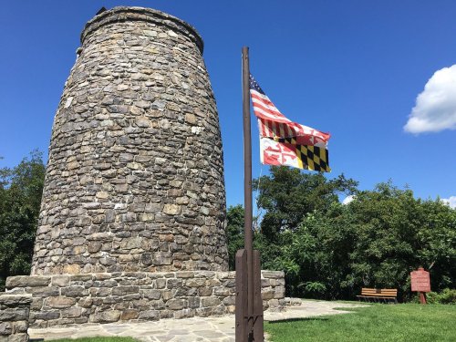 Stone tower with American and Maryland flags, surrounded by greenery and a clear blue sky.