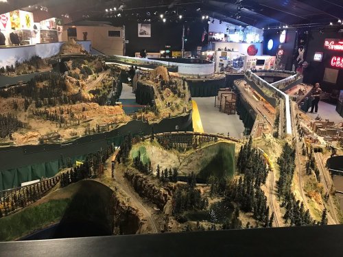 A large model train layout featuring mountains, forests, and tracks, with people observing in a dimly lit exhibition space.