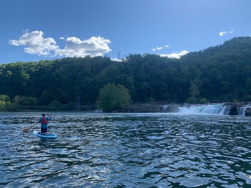 A person paddleboarding on a calm lake with a waterfall and lush green hills in the background under a blue sky.