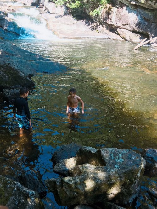 Two boys play in a clear, shallow stream surrounded by rocks and greenery, with a waterfall in the background.