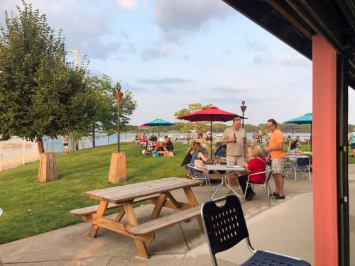 People gather at a lakeside park with picnic tables and umbrellas, enjoying a sunny day by the water.