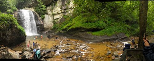 A panoramic view of a waterfall surrounded by lush greenery, with people exploring the rocky area and wading in the water.