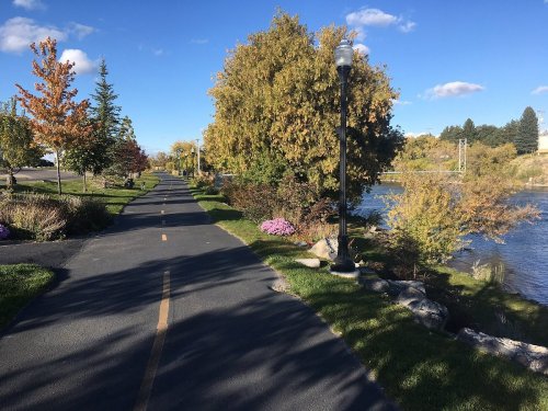 A scenic pathway lined with trees and flowers, alongside a river under a clear blue sky.