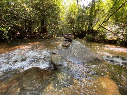 A serene stream flows over rocks, with a small stack of stones balanced on a larger rock in the sunlight.